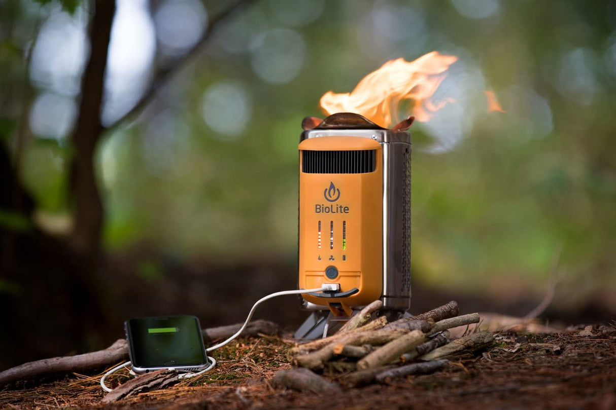 The BioLite CampStove 2 outside laid standing up on the ground outdoors with bundles of sticks surrounding it and charging a mobile device via white usb cable. On the campstove are lit up indicators of the different energy sources.