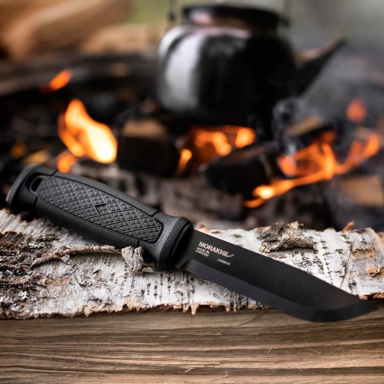 Morakniv Garberg Carbon Survival Knife laying on log next to a campfire. 