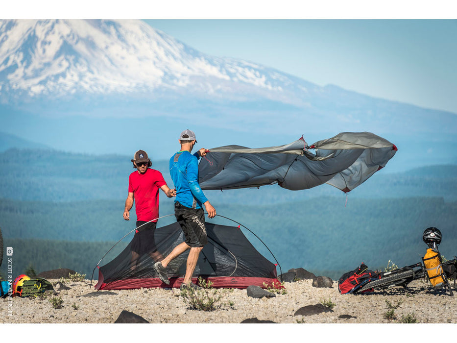 2 guys setting up the MSR Carbon Reflex Ultralight Tent. A man wearing a blue shirt is placing the weather cover over the tent while another man wearing a red shirt helps. Two bicycles are shown on the ground and a large mountain in the background.