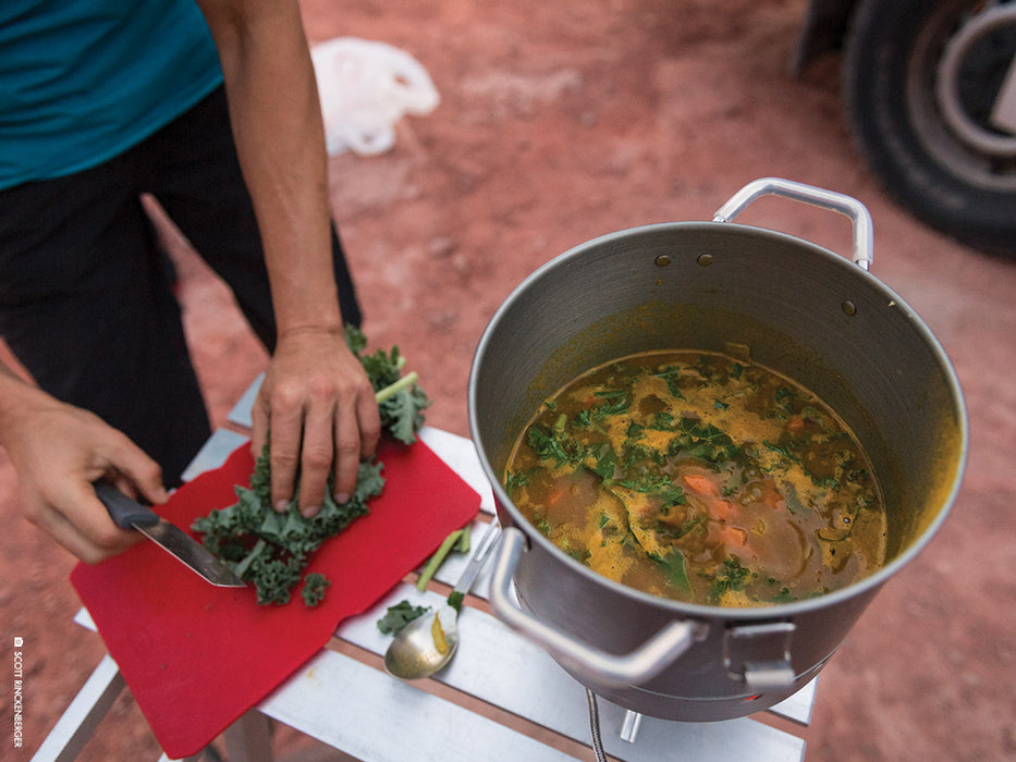 A person wearing a blue shirt and black pants cutting up kale and throwing it in to the MSR WindBurner Large Stock Pot. In the pot is a soup stock with carrots and beef. The stock pot is laid on a foldout aluminum camping table. Red dirt populates the background with a tire of a vehicle. 