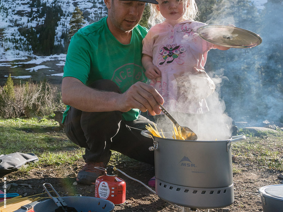 A father in a kelly green shirt turns spaghetti pasta in a Large WindBurner Stock pot while his blonde haired daughter wearing a pink dress holding the pot lid. Steam rises from the pot as they cook the pasta together on a camp stove. A snow covered hill is in the background.