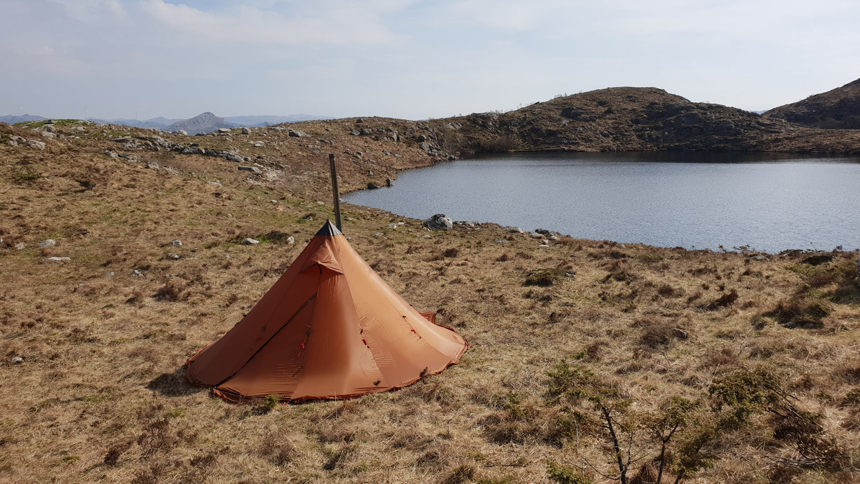 A copper coloured NorTent Iavvo 4 season tent beside a lake in the badlands. Rocks and brown grass populate the surroundings.