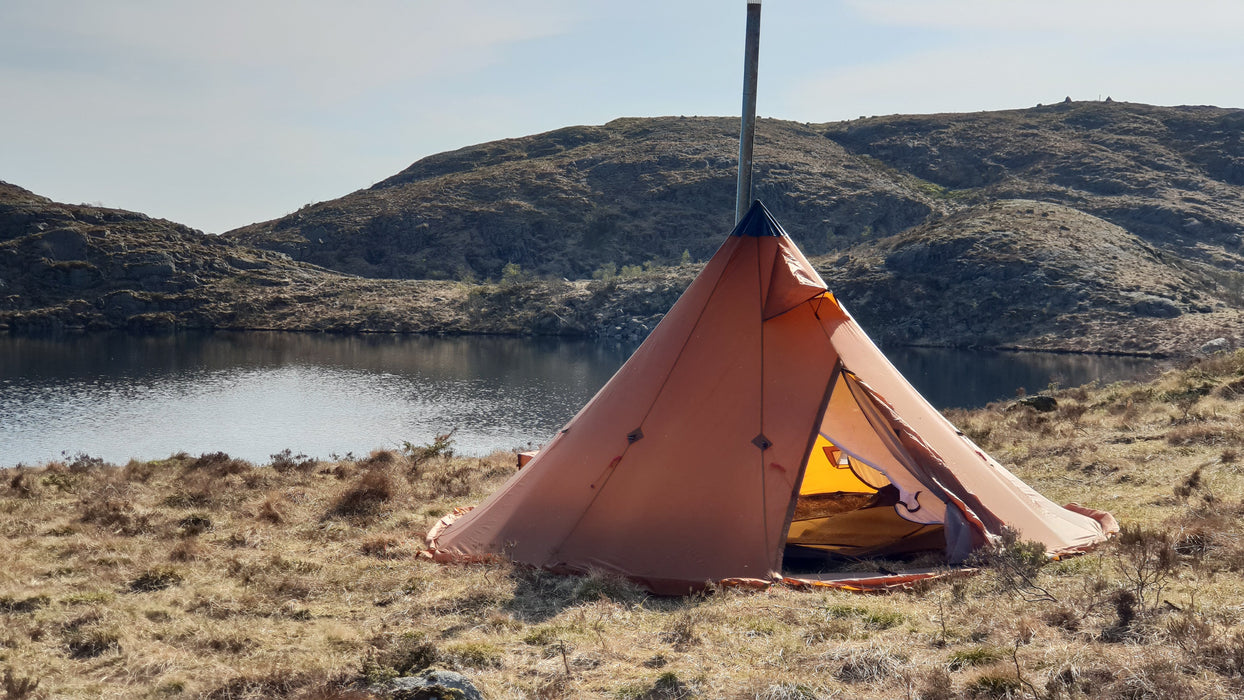 The Iavvo 4 winer hot tent set up near a lake on a hillside on a hot summer day. The grass on the ground around it is a brown colour with small bushes scattered and in the distance is a large hillside across the water.