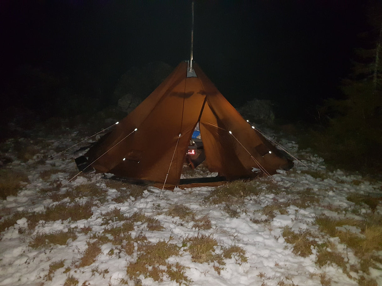 Inside the entry way of a NorTent Iavvo 6, you can see a mans foot and a small stove burning wood to heat up the inside of the tent. Surrounding the tent is snow and grass shrubs.