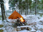 A fire pit is lit beside a NorTent Iavvo 6 winter hot tent. The tent is a brown copper colour and the fire infront of it is large with tree branches from the surrounding area. Spruce trees and a snow covered ground populate the area.