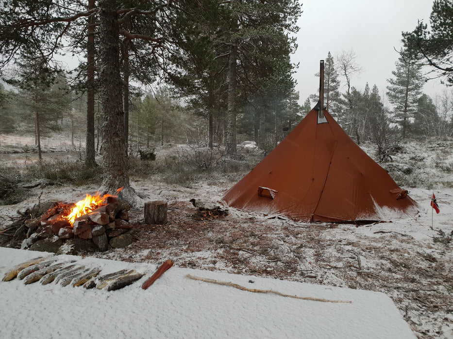Fire logs are laid out on a table, in the background is a large fire pit surrounded by tree stumps to contain the flames. A Nortent Iavvo 6 Winter Hot tent is beside the fire to the right with the chimney erect in a brown color.