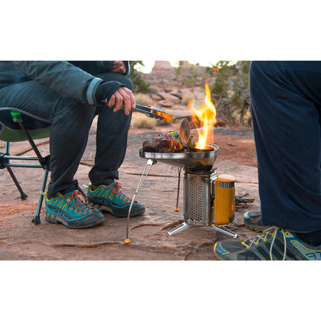 2 hikers huddling over the grill top of BioLite CampStove 2 cooking steaks with vegetables on top. The hikers are wearing bright coloured hiking shoes on top of a rock face with a desert looking background view.