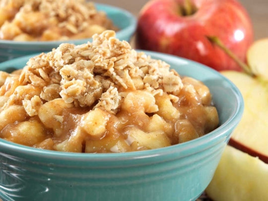 A teal bowl of Mountain House Apple Crisp prepared freeze dried food. Apples and another bowl are faded in the background. 