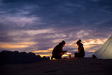 A couple sitting around a fire made by BioLite CampStove 2 at sunset outside of their tent.