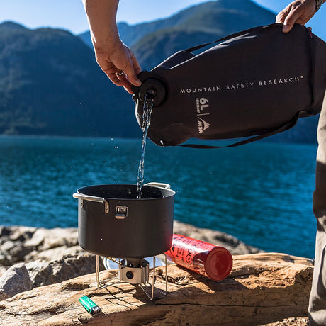 A person pouring filtered water into a camp stove pot from a Mountain Safety Research (MSR) rugged water storage bag. A large lake and hillside are shown in the distance. 