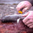 A person filleting a fish with a Morakniv Companion knife. The fish is laid upond a tree log and the fishermans hands are covered in dirt and filth.