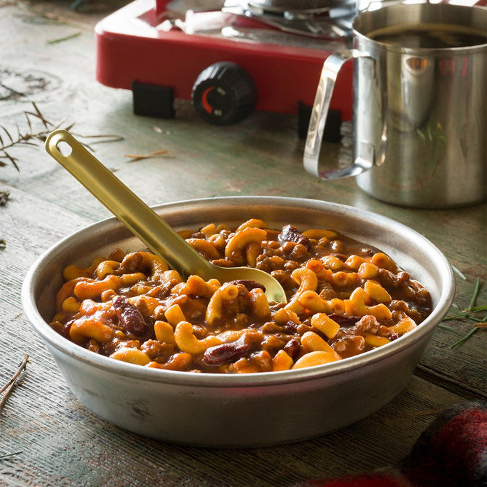 A medium sized camping bowl full of the prepared Mountain House Chili Mac with Beef. A camping spoon is placed in the bowl and in the background is a stainless steel mug and a red hot plate. The items are placed on a park table with fallen spruce needles around them.