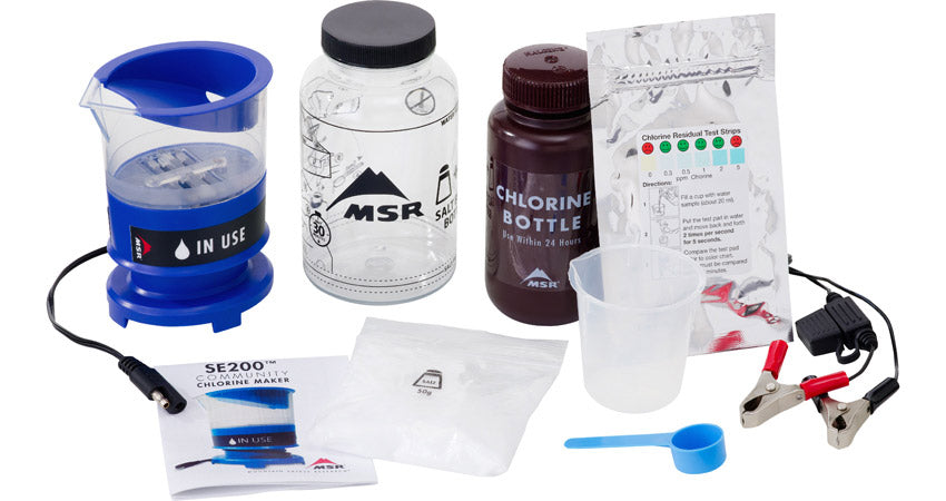Mountain Safety Research Purification Kit with the chlorine bottle in black to the right, the clear water bottle to the left and the purifying chamber to the far left. A relfective mylar bag of chlorine tablets is shown alongside a pair of black and red leads for jumpstarting. A Blue teaspoon measurer is shown beside a larger clear measuring cup. 