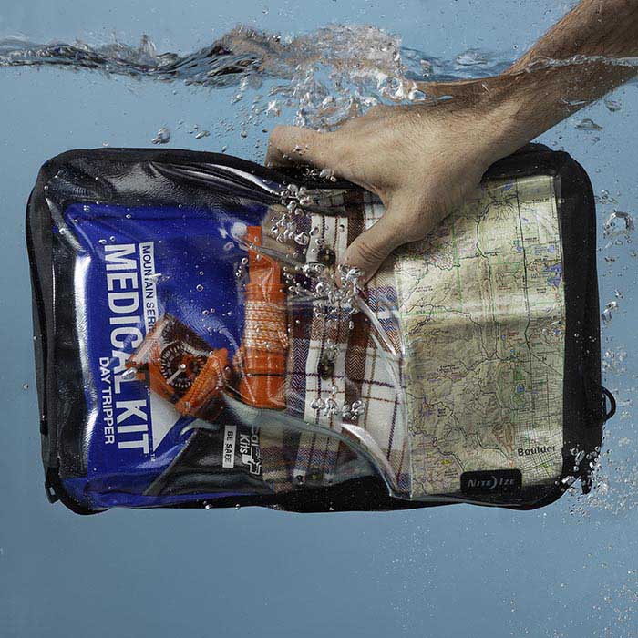 A person holding a Nite-ize waterproof packing cube submerged in water with a light blue background. In the packing cube is a medical kit, compass, map and change of clothes.