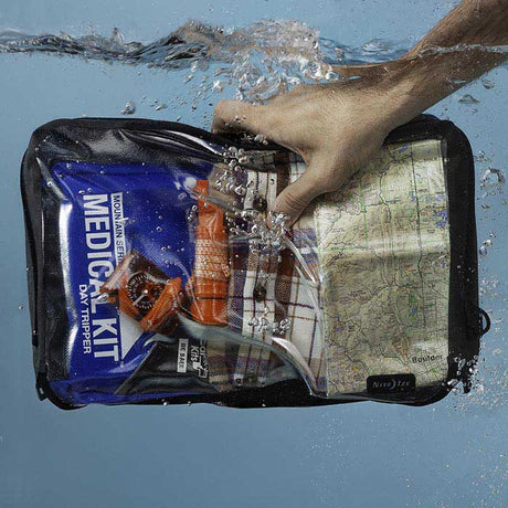 A person holding a Nite-ize waterproof packing cube submerged in water with a light blue background. In the packing cube is a medical kit, compass, map and change of clothes.