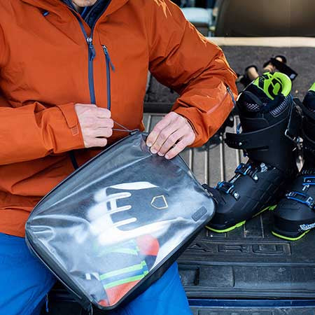 A man wearing an orange jacket closing up a bag of ski gloves. The bag is a Nite-ize packing cube and is also see-through. His skii boots are shown in black and lime green on the back of his suv.