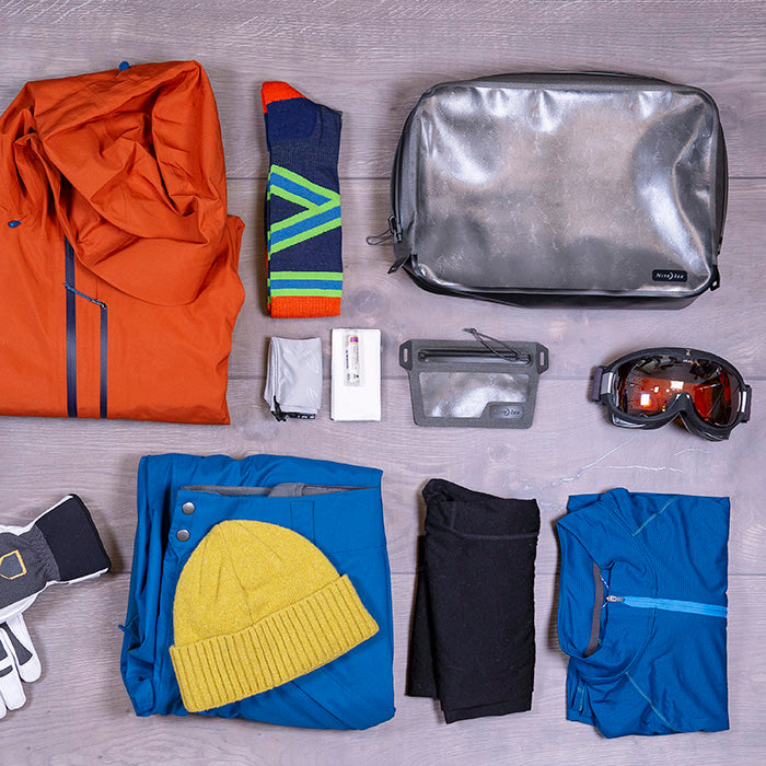 Outdoor clothing organized on a hardwood floor near a Nite-ize packing cube. There are snow goggles, an orange jacket, blue snow pants, yellow toque, blue sweater, black thermal tights, blue and lime green socks, white and grey gloves and a smaller waterproof satchel for wallet contents.
