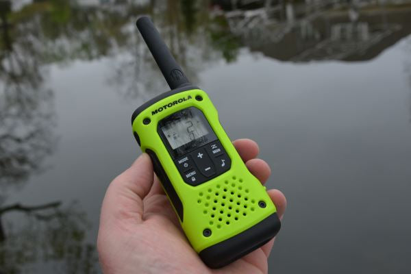 A person holding a T600 Two way Radio in one hand. The radio is a lime green with black accents and buttons. In the background is a lake with a reflection of a house in it.