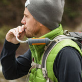 A man wearing a grey toque and a lime green vest is showing drinking from a Muv Eclipse water filter hose from his hydration pack.