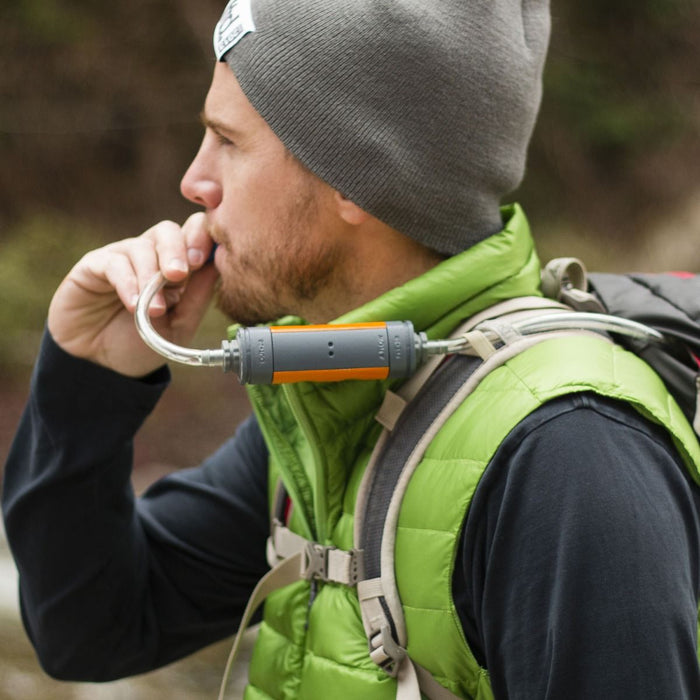 A man wearing a grey toque and a lime green vest is showing drinking from a Muv Eclipse water filter hose from his hydration pack.