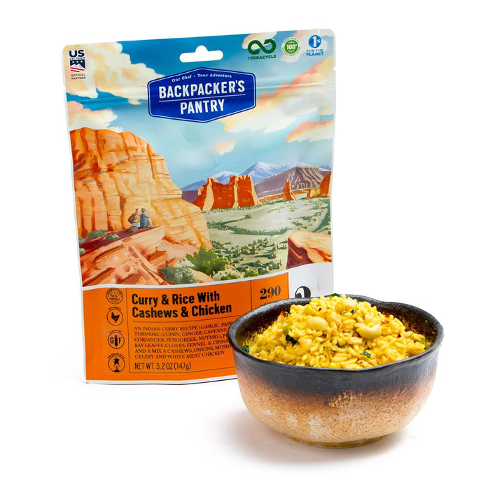 bowl of cashew curry rice with chicken alongside the backpackers pantry freeze dried bag. Drawing of 2 people enjoying the beautiful site of a canyon and mountains, during a day hike.