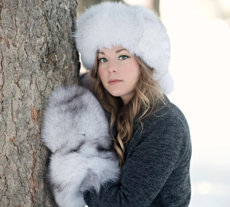 Woman wearing the Blue Fox Fur Mitts with a Blue Fox Fur hat up against a tree in the outdoors with snow in the background.