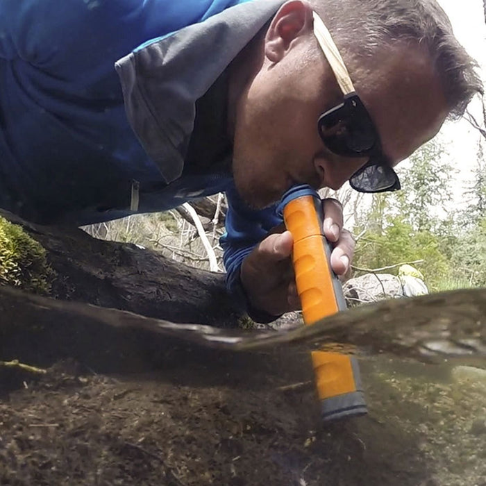 A man wearing sunglasess and a blue coat is shown drinking water from a river using the MUV eclipse water filter. The filter is in orange and grey with finger grooves up the body.