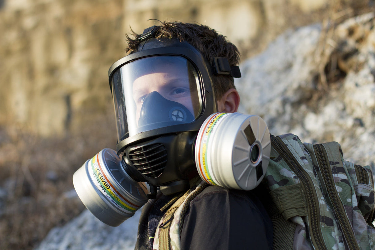 A man wearing the CM-6M TACTICAL GAS MASK with two repirator filters showing. On his is back is a army camouflaged backpack and a cliffside shows in the background.