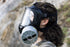 A woman with long brunette hair wearing the CM-6M TACTICAL GAS MASK with a large repirator filter shown. In the background is a cliffside and the woman is staring out in the distance.