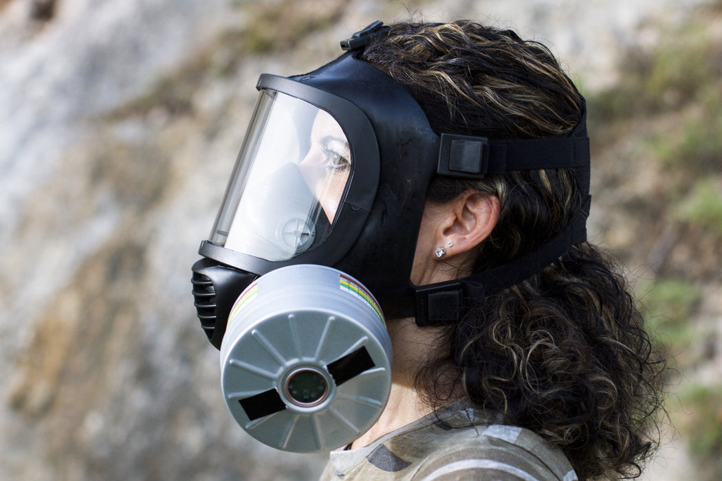 A woman with long brunette hair wearing the CM-6M TACTICAL GAS MASK with a large repirator filter shown. In the background is a cliffside and the woman is staring out in the distance.