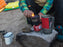 Someone sparking a camp stove with a lighter. A sleeping bag is in the background in yellow, 3 camping mugs sit by the stove. 