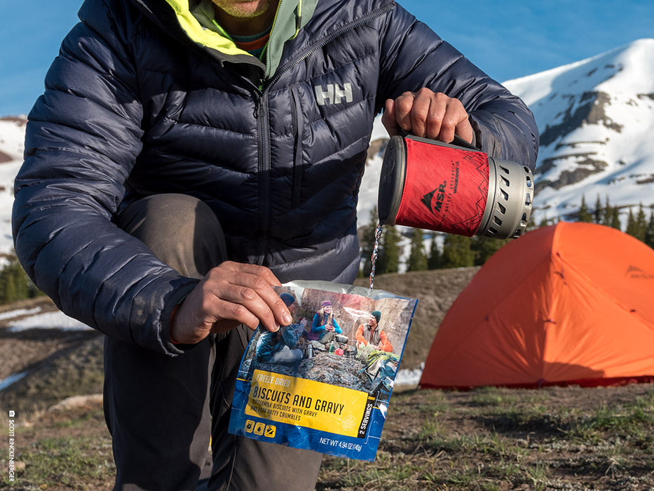 Man in a Helly Hansen Black puffer jacket pouring boiled water out fo a MSR Windburner pot into a freeze dried food package of biscuits and gravy from Moutnain House. A Orange MSR Tent is in the background with A snow covered mountain top and spruce tree's.