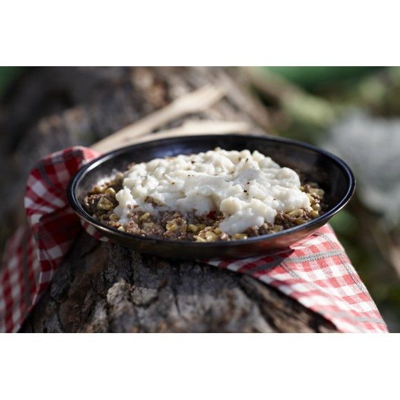 A bowl of prepared freeze dried Shepherds Pie on a red and white plaid coloured napkin resting on a tree log.