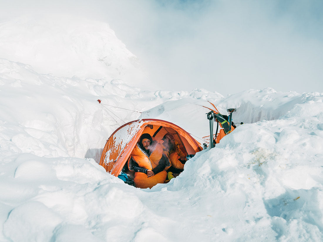2 men camped out in a tent wearing Polar Ranger sleeping bag suits. A man's breath is visible in the cold weather and snow completely surrounds them.