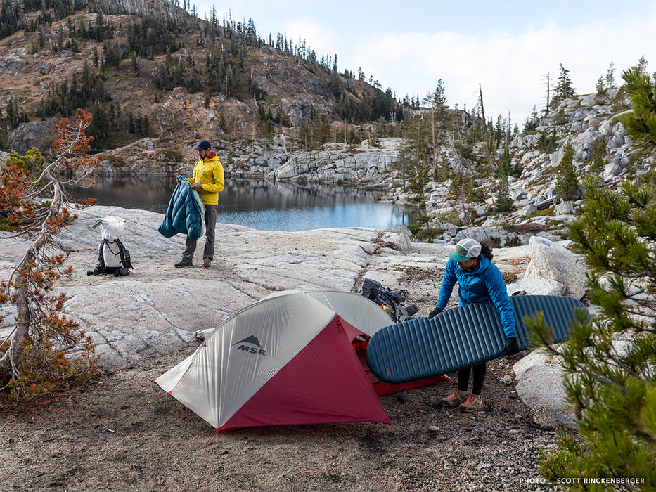 A person in a blue jacket placing a Neoair Uberlite mattress in their MSR two person tent camped out infront of a calm lake. A person wearing a yellow jacket unravels his blue sleeping bag in the distance.