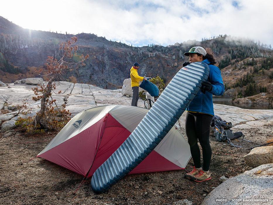 A person blowing air into a Thermarest Neoair mattress beside a red and grey coloured tent. The person is wearing running tights and a blue jacket. A person wearing a yellow jacket is unpacking their gear in the background. A hillside with scattered spruce trees and clouds is shown.