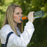 A woman drinking from a water bottle using the Muv Survivalist water filter attached to it. The woman is wearing a white coat with black stripe on the shoulder.